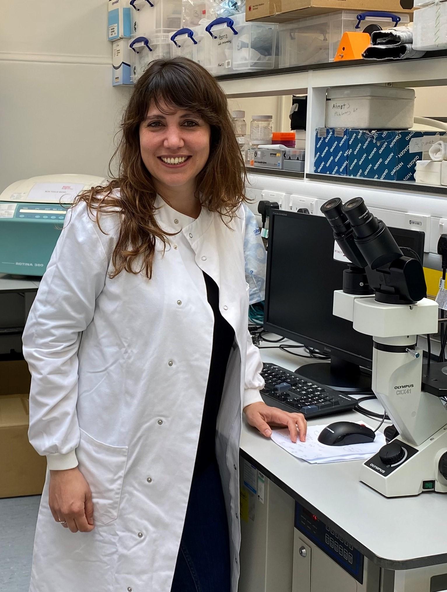 Ana Rio-Machin wearing a white lab coat, standing in the laboratory next to a microscope.