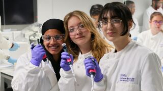 Three pupils stand smiling in the BCI's laboratories, wearing labcoats and goggles and holding pipettes.