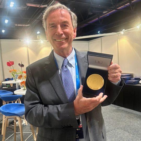 John Gribben stands smiling at the viewer, holding a gold medal in a box.