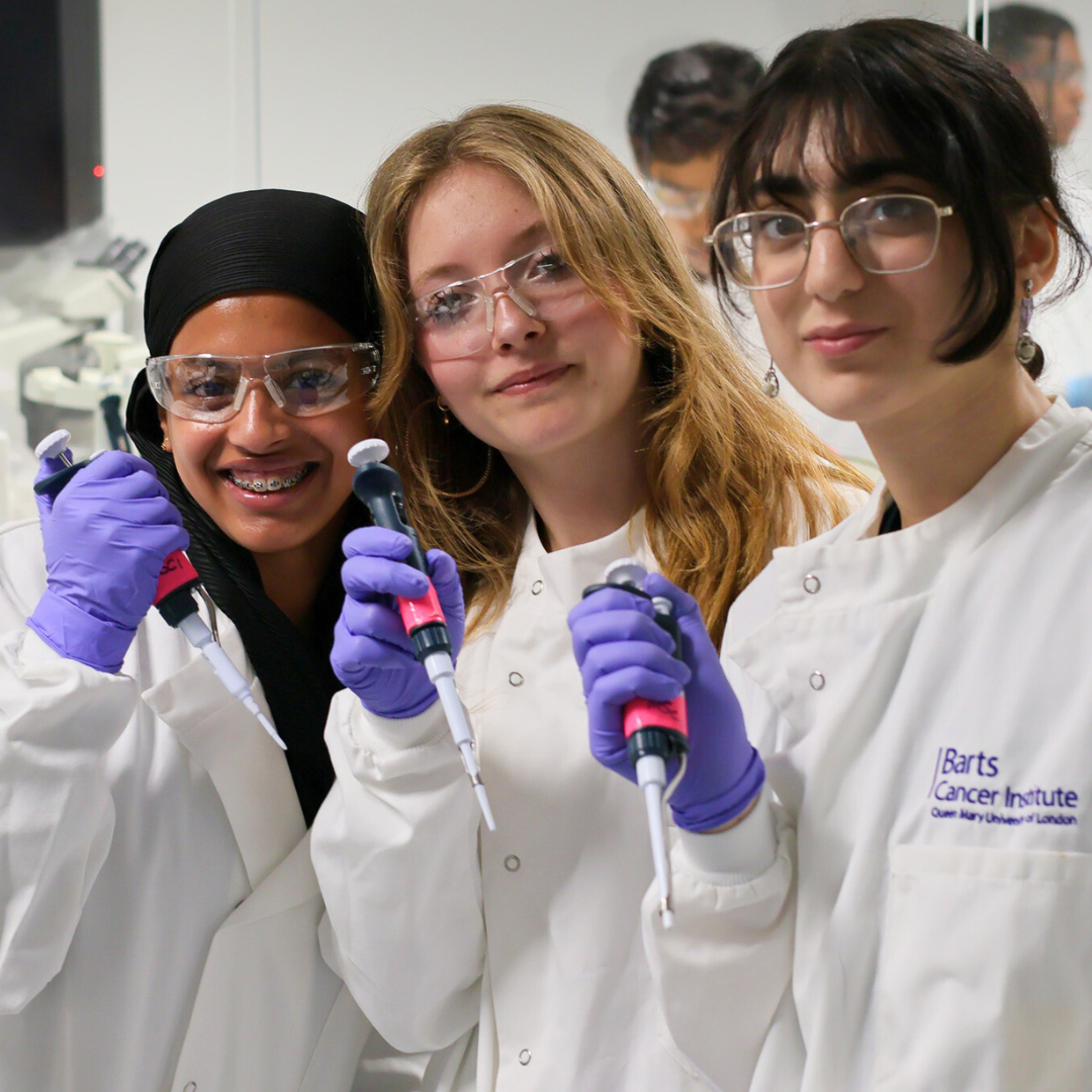 Three pupils stand smiling in the BCI's laboratories, wearing labcoats and goggles and holding pipettes.